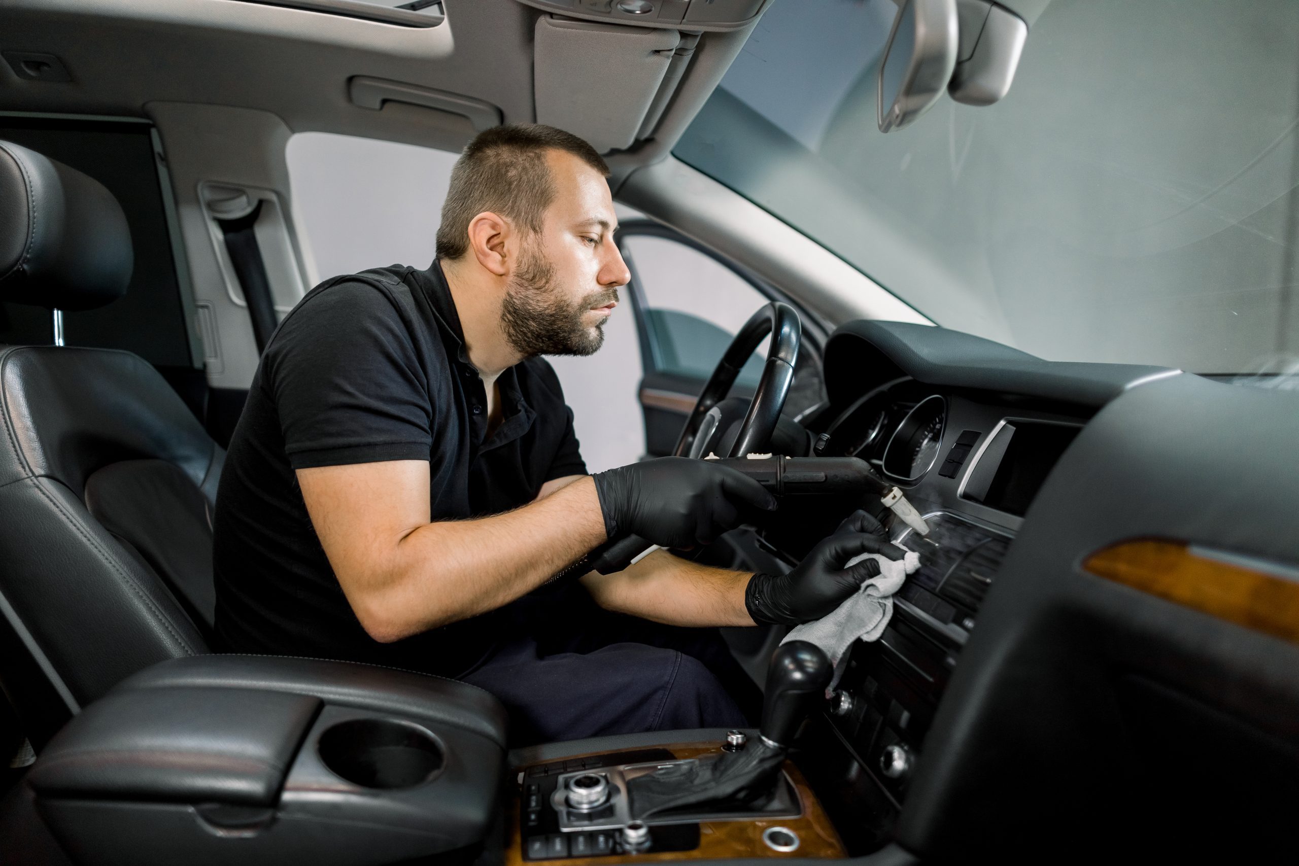 Service cleaning of air conditioner of the car with hot steam. Concentrated professional Caucasian male worker in black uniform, sitting in front seat of modern car and using hot steam cleaner Service cleaning of air conditioner of the car with hot steam. Concentrated professional Caucasian male worker in black uniform, sitting in front seat of modern car and using hot steam cleaner.